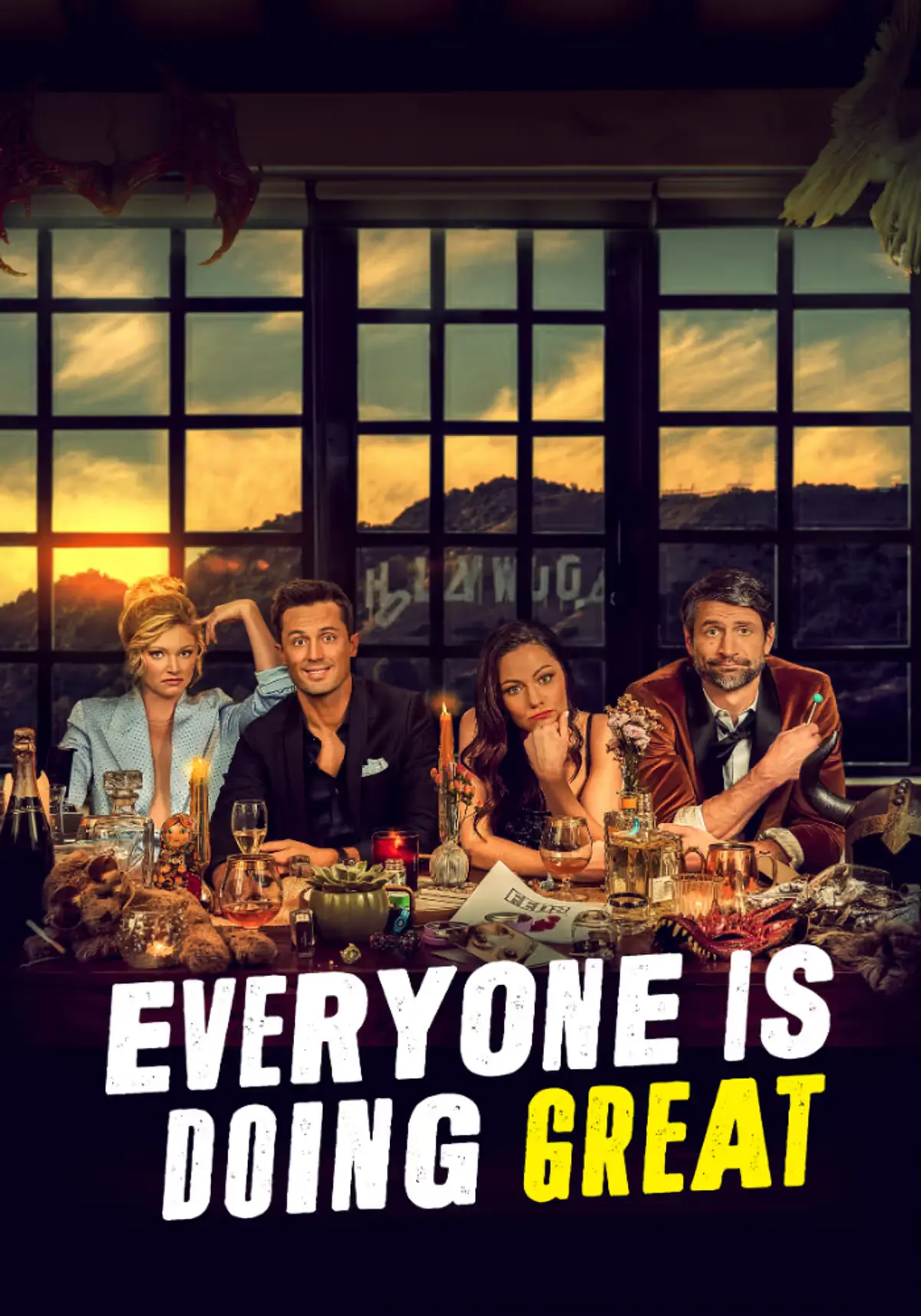 Cast of Everyone Is Doing Great seated around a cluttered dinner table with drinks, candles and props, posed in front of a large window at sunset, with the show’s title displayed across the bottom.